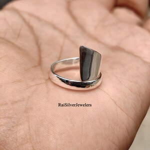 Può includere: Un anello in argento lucido caratterizzato da un design aperto con un elemento decorativo verticale piatto e rettangolare. La fascia è liscia e arrotondata, e il testo RaiSilverJewelers è stampato in nero vicino alla base dell'anello sulla pelle.