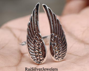 Peut inclure: Une bague argentée composée de deux éléments symétriques en forme d'ailes qui se rejoignent au centre. La surface métallique présente une finition texturée et oxydée soulignant les détails des plumes. Le texte RaiSilverJewelers est visible.