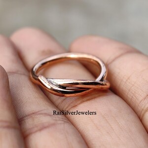 May include: A polished copper-toned ring featuring a bypass design where the band ends overlap. The metal has a warm, metallic sheen and a smooth, rounded surface. The text RaiSilverJewelers is printed in black on the surface of the ring.