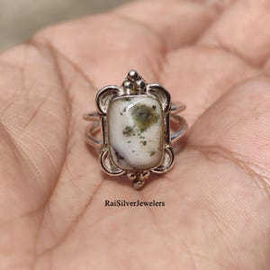 May include: A silver ring featuring a rectangular white stone with dark green inclusions. The setting has a decorative frame with small silver spheres at the top and bottom. The text RaiSilverJewelers is printed at the bottom of the image.