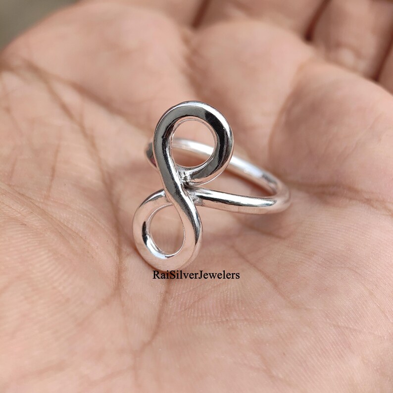 Peut inclure: Une bague en argent poli pr&eacute;sentant un motif de symbole infini vertical. La surface m&eacute;tallique est lisse et r&eacute;fl&eacute;chissante, formant une boucle continue. Le texte RaiSilverJewelers est imprim&eacute; en petites lettres noires &agrave; la base de la bague.