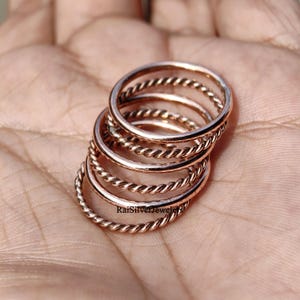 Op de afbeelding: Een set van vijf gestapelde roségouden ringen, met een mix van glad gepolijste banden en gedraaide touwstructuur banden. De ringen zijn in een geneste formatie gerangschikt en de tekst RaiSilverJewelers is zichtbaar op het oppervlak van de onderste ring.