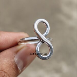 Peut inclure: Une bague en argent poli pr&eacute;sentant un motif de symbole infini vertical. La surface m&eacute;tallique est lisse et r&eacute;fl&eacute;chissante. Le texte RaiSilverJewelers est imprim&eacute; en petites lettres noires pr&egrave;s de la base de la boucle infinie sur l'anneau.