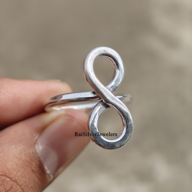 Peut inclure: Une bague en argent poli pr&eacute;sentant un motif de symbole infini vertical. La surface m&eacute;tallique est lisse et r&eacute;fl&eacute;chissante. Le texte RaiSilverJewelers est imprim&eacute; en petites lettres noires pr&egrave;s de la base de la boucle infinie sur l'anneau.