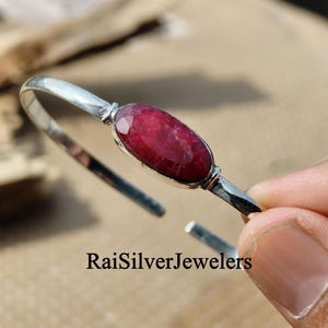 May include: A silver cuff bracelet featuring a central oval-shaped, faceted deep red gemstone. The metal band is polished and smooth, with the text RaiSilverJewelers visible at the bottom of the image. The design is open-ended for adjustment.
