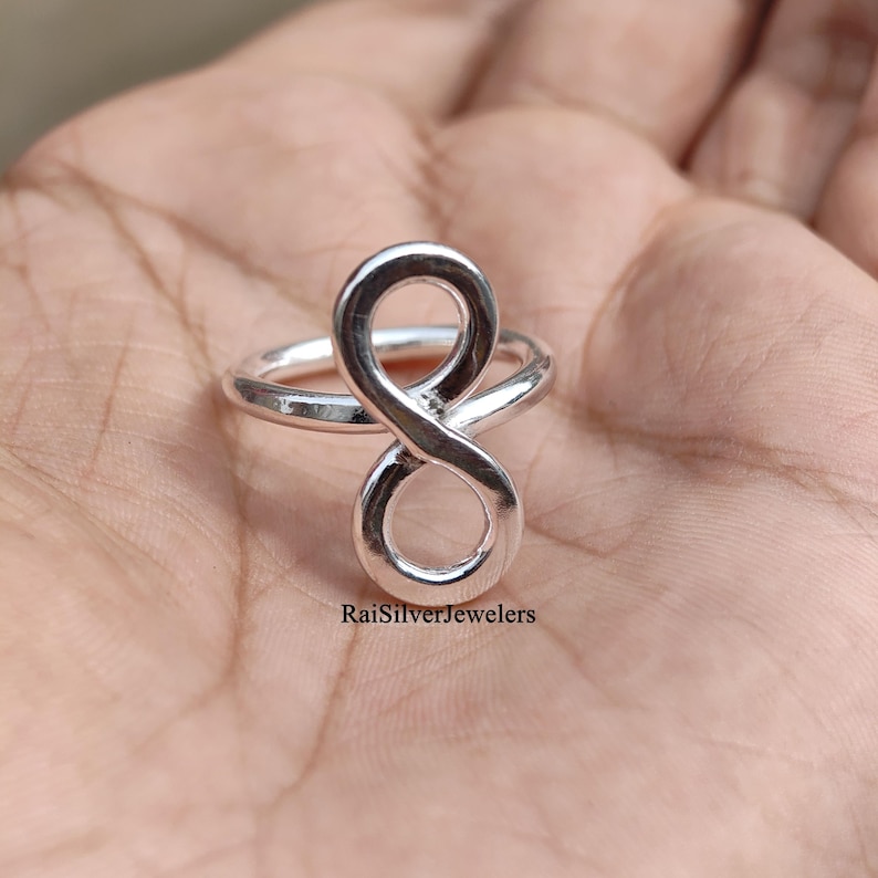 Peut inclure: Une bague en argent poli pr&eacute;sentant un symbole de l'infini vertical centr&eacute; sur l'anneau. Le m&eacute;tal a une surface lisse et r&eacute;fl&eacute;chissante. Le texte RaiSilverJewelers est imprim&eacute; en petites lettres noires pr&egrave;s de la base du symbole.