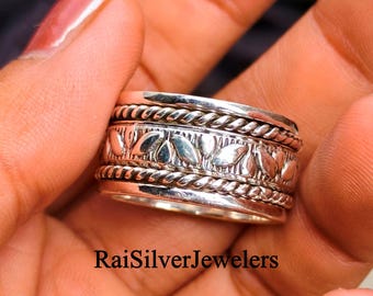 Peut inclure: Une bague en argent présentant une bande centrale avec un motif de feuilles répétitif, encadrée par deux bandes torsadées en forme de corde sur les bords supérieur et inférieur. Le métal a une finition polie avec des accents oxydés sombres. Texte : RaiSilverJewelers.