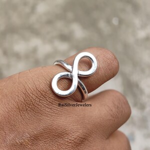 Peut inclure: Une bague en argent poli pr&eacute;sentant un symbole de l'infini bien visible sur le devant. L'anneau en m&eacute;tal est lisse et r&eacute;fl&eacute;chissant, formant une boucle continue. Le texte RaiSilverJewelers est visible sur la partie inf&eacute;rieure de la bague.