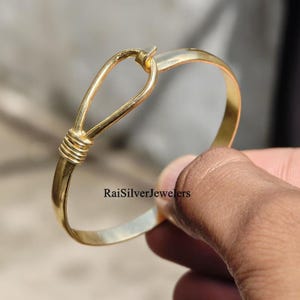 May include: A gold-toned metal bangle bracelet featuring a hook-and-loop style closure with a decorative coiled wire detail at the junction. The surface of the bracelet is smooth and polished. The text RaiSilverJewelers is printed on the metal.