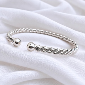 Puede incluir: Una pulsera tipo brazalete de color plateado con una textura trenzada similar a una cuerda a lo largo de la banda y una esfera lisa y pulida en cada uno de los dos extremos abiertos. El metal tiene un acabado metálico reflectante.