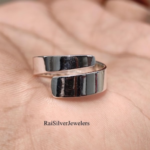 Peut inclure: Une bague bypass en métal argenté poli présentant une surface lisse et réfléchissante avec un design ouvert. L'anneau métallique se courbe pour créer deux extrémités qui se chevauchent. Le texte RaiSilverJewelers est visible en bas.