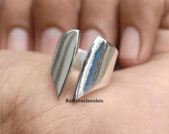 Peut inclure: Une bague en argent poli présentant un design ouvert de style bypass avec deux extrémités distinctes, inclinées et effilées. La surface du métal est lisse et réfléchissante. Le texte RaiSilverJewelers est imprimé en petites lettres noires à la base.
