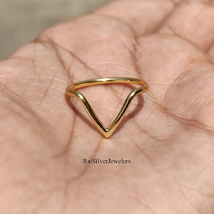 May include: A gold-coloured V-shaped ring with a smooth, polished finish. The band features a distinct chevron or pointed design that centres to a point. The text RaiSilverJewelers is printed in black at the bottom of the image area.