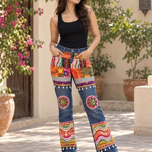 Puede incluir: Una camiseta sin mangas negra combinada con pantalones vaqueros azules que presentan paneles de patchwork bordados en colores vibrantes, detalles de espejos circulares y conchas de cauri blancas colgando del borde horizontal.