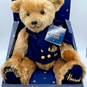 Pode incluir: Um urso de peluche castanho com um colete de veludo azul-marinho com botões dourados, sentado numa caixa azul-escura com estrelas douradas. O urso tem Millennium 2000 bordado nas patas e uma etiqueta Harrods 2000 no pescoço.