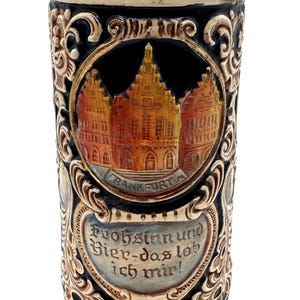 Peut inclure: Une chope à bière cylindrique en céramique avec un bord et une base de couleur beige. Le corps noir présente un relief coloré de bâtiments de Francfort dans un cadre circulaire, avec le texte "Frankfurt" et "Frohsinn und Bier-das lob ich mir!".