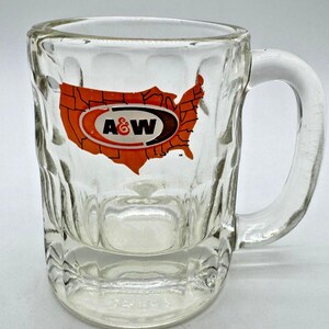 May include: A clear glass mug with a thick handle and a dimpled exterior texture. The front features an orange graphic of the United States map with the black and white A&W logo centered over the map. The glass has a heavy base and a smooth rim.