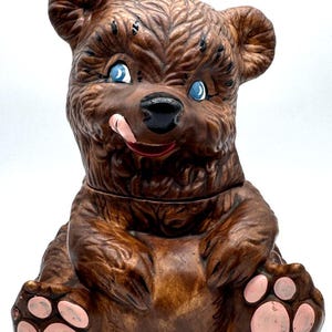 May include: A brown ceramic bear figurine sitting in an upright position. The bear has textured fur, bright blue eyes, a black nose, and a small pink tongue sticking out. The paws feature light pink pads on the soles, and the overall finish is glossy.