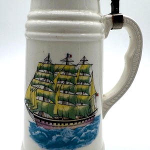 Peut inclure: Une chope à bière en céramique blanche ornée d'une illustration colorée d'un voilier à plusieurs mâts aux voiles vertes sur des vagues bleues. La chope possède un corps cylindrique haut, une anse courbée et un couvercle métallique décoratif.