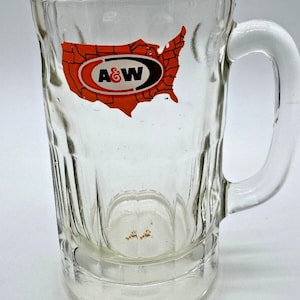 May include: A clear glass beer mug featuring a vertical ribbed texture and a sturdy handle. The front of the mug displays a graphic of the United States in orange with black state outlines, overlaid with the A&W logo in black, white, and red.