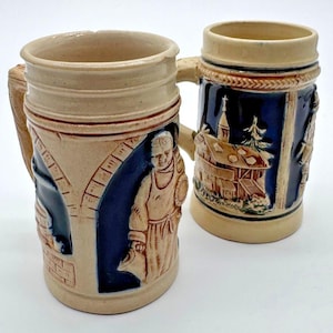 May include: Two ceramic beer steins featuring a tan and dark blue colour scheme. The left stein displays a relief of a person in an archway, while the right stein depicts a building with a steeple. Both mugs have a handle and a cylindrical shape.