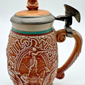 May include: A ceramic beer stein featuring a relief design with the text WILD WEST. The body depicts a figure on horseback with a rifle, accented in tan, white, and teal. It includes a metal hinged lid topped with a small sculpted figure.