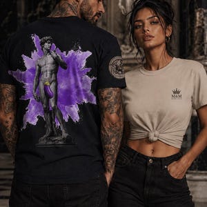 May include: A black t-shirt featuring a graphic of a statue with a purple aubergine overlay against a purple splash, and a beige t-shirt with a small crown logo and the text M&M. Both shirts are paired with black denim jeans.