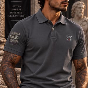 Può includere: Una polo a maniche corte color grigio antracite con un logo a corona bianco e la scritta M&M CLOTHING sul petto sinistro, e un testo sulla manica destra che recita THE WORLD SAW THE SNAKE HISTORY FORGOT THE WOMAN in un layout a caratteri sovrapposti.