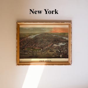 Op de afbeelding: Een ingelijste vintage-stijl print van een stadsplattegrond van New York met een vogelvluchtperspectief van de haven met schepen en gebouwen. De tekst New York staat boven de lijst en onderaan de print. De lijst heeft een verweerde gouden kleur.