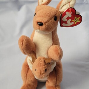 May include: A plush kangaroo toy with light brown fur and a cream-coloured belly. The kangaroo has a small joey in its pouch and a heart-shaped red and yellow Ty Beanie Baby tag attached to its ear. The toy features black eyes and a black nose.