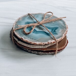 Peut inclure: Ensemble de quatre sous-verres en agate li&eacute;s ensemble avec de la ficelle. Les sous-verres sont de couleur bleu clair avec des bandes brunes et blanches.