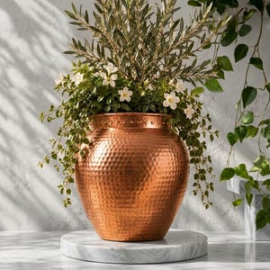 Peut inclure: Un grand pot de fleurs en cuivre martelé, de forme arrondie avec une large ouverture et une surface métallique texturée. Le pot est rempli d'un arrangement luxuriant de feuillage vert, incluant des feuilles fines et des petites fleurs blanches.