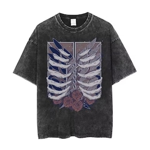 May include: A charcoal grey acid-wash t-shirt featuring a graphic print of a ribcage structure layered over two blue and grey wings. Below the ribcage are several dark red roses. The design is framed by a thin white rectangular border.