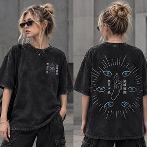 Puede incluir: Una camiseta gris carbón con efecto lavado al ácido, con un pequeño gráfico de un ojo y texto en japonés en el pecho izquierdo. La parte trasera muestra un gran diseño circular con varios ojos azules, una mano y texto en japonés blanco.