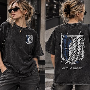 Può includere: Una t-shirt grigio antracite con effetto lavaggio acido, caratterizzata da una piccola grafica sul petto e un grande disegno sul retro. Il design posteriore mostra uno scudo con ali blu e bianche e la scritta "WINGS OF FREEDOM".