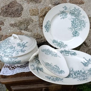 May include: A collection of cream-colored ceramic dinnerware featuring a teal floral pattern. The set includes a lidded tureen with side handles, a large round dinner plate, a smaller oval serving dish, and a large oval serving platter.