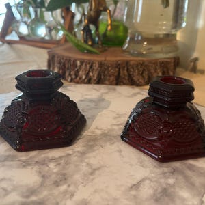 May include: Two deep red glass candle holders featuring a hexagonal base and a raised geometric pattern. Each piece has a central opening for a taper candle and a wide, stable bottom with intricate embossed detailing across the exterior surface.