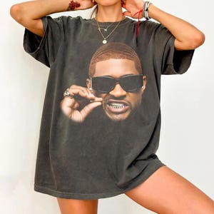 May include: A charcoal grey short-sleeve t-shirt featuring a large, centred graphic print of a man with short hair, wearing dark sunglasses, and displaying silver teeth grills. The man is holding a small object near his mouth with his right hand.