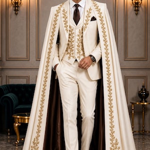 May include: A cream-colored three-piece suit consisting of a vest, trousers, and jacket, paired with a long matching cape. The vest and cape feature intricate gold embroidery in a leaf pattern. A dark brown tie and dark brown dress shoes complete the look.