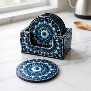 May include: A set of round drink coasters featuring a dark blue, light blue, and white dot-painted mandala pattern with a central eye motif. The coasters are stored in a matching rectangular holder with the same intricate dot-painted design.