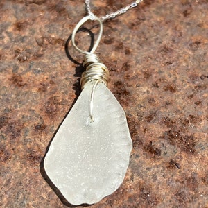 May include: A handcrafted necklace featuring a piece of frosted sea glass, wrapped in silver wire, suspended from a delicate silver chain. The sea glass has a smooth, irregular shape and a translucent, frosted appearance. The necklace is a unique, beach-inspired accessory.