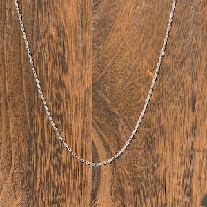 May include: A delicate silver necklace with a fine chain. The necklace is displayed against a warm, wooden background. The chain is composed of small, interconnected links.