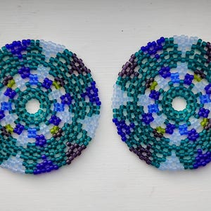 May include: Two circular beaded coasters featuring a geometric pattern in shades of teal, royal blue, dark purple, and translucent light blue. Each coaster has a small circular opening in the centre and is constructed from small, uniform glass beads.