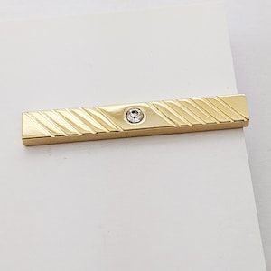 Könnte beinhalten: Eine goldfarbene Krawattenklammer mit diagonal gerilltem Muster und einem kleinen, runden, klaren Edelstein in der Mitte. Der rechteckige Stab hat eine polierte Oberfläche.