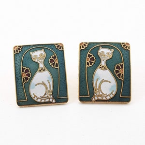 May include: A pair of square earrings with a teal background and a gold border. Each earring features a white cat design with gold accents and maroon flowers. The earrings have a vintage aesthetic.