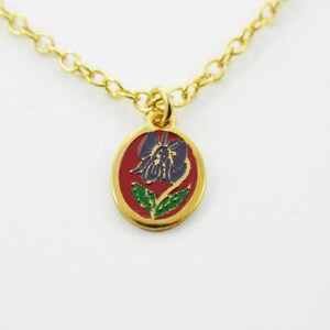 Può includere: Collana dorata con un ciondolo con un motivo floreale. Il ciondolo ha uno sfondo rosso con un fiore blu e viola, foglie verdi e un bordo dorato. La catena è in stile a maglie delicate.