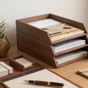 Può includere: Un vassoio portadocumenti in legno scuro a tre livelli contenente fogli e buste, accompagnato da un portapenne in legno abbinato, un piccolo portacarte in legno, una pila di biglietti con scritto Confidential, una penna e un vaso dorato.