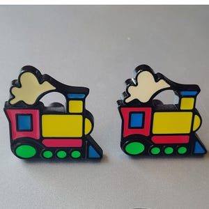 May include: A pair of enamel pins shaped like steam locomotives. Each train features a black outline with a yellow body, red accents, a blue window, a green wheel section, and a cream-colored puff of smoke rising from the top chimney stack.