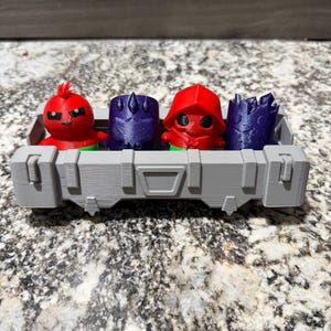 May include: A gray rectangular 3D-printed crate containing four small figurines. The figures include two red characters with green bases and two dark purple textured characters. The crate features a structured design with vertical panels.