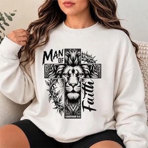 May include: A cream-colored crewneck sweatshirt featuring a black graphic print of a lion's face centered within a cross. The design includes a crown of thorns, vines, and the text "MAN OF FAITH" and "I CORINTHIANS 16:13" in a bold font.